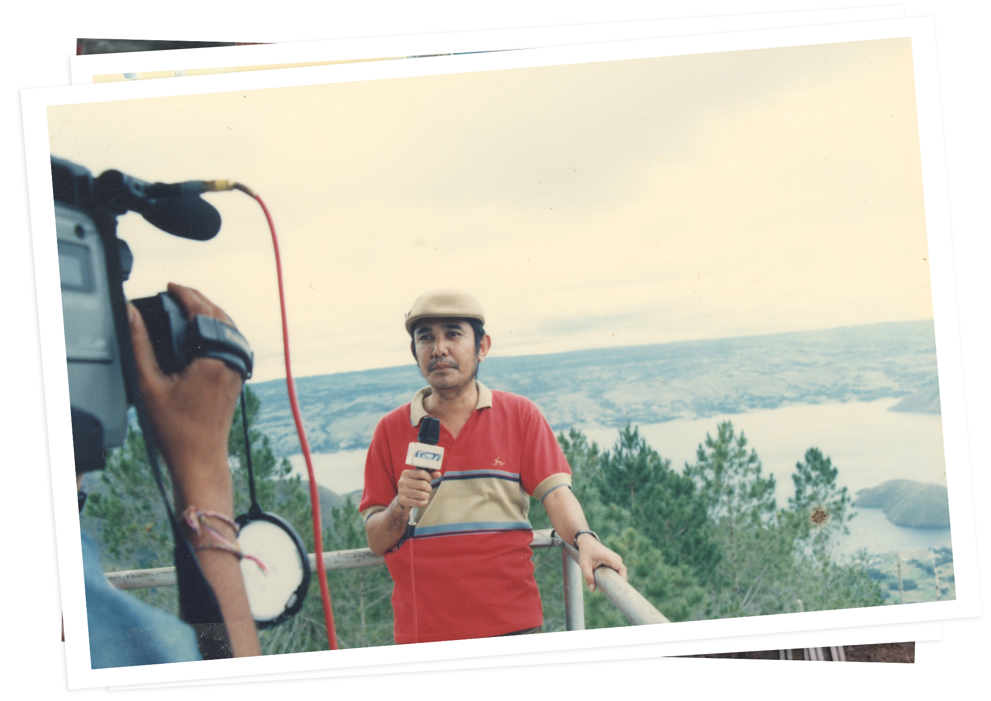 Ersa Siregar, wearing a cap and holding a microphone, reports outdoors with hills behind him.